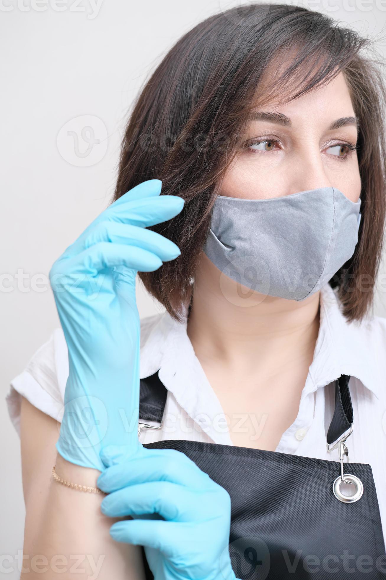 donna che indossa un grembiule che indossa guanti protettivi blu. donna  bruna con maschera tessile su un viso che si prepara a fare la procedura di  bellezza. estetista per la depilazione. 8250911