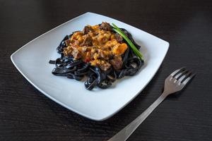 pasta nera al nero di seppia. pasta di semola di grano duro al nero di seppia con salsa di manzo e peperoni su un piatto quadrato decorato con cipolle verdi con forchetta foto