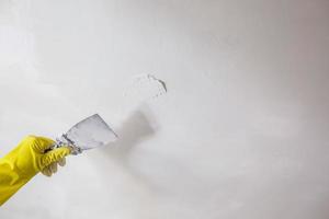 la mano del lavoratore in guanti gialli che tiene una spatola che rattoppa un foro con una spatola con gesso o mastice nel muro bianco. processo di ristrutturazione e riparazione, rimodellamento interno della stanza in condominio foto