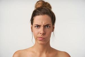 perplessa giovane donna attraente con alta acconciatura chignon in piedi su sfondo bianco, guardando alla telecamera con faccia seria e accigliata foto