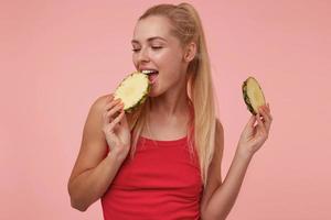 foto interna di una bella giovane donna bionda in camicia rossa in posa su sfondo rosa con gli occhi chiusi, godendo del gusto dell'ananas fresco