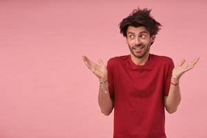 giovane uomo dai capelli scuri con la barba in piedi su sfondo rosa in maglietta rossa, guardando da parte con la faccia perplessa e alzando i palmi delle mani, contraendo la fronte e alzando le sopracciglia foto