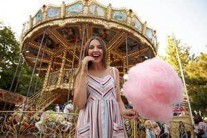 Ritratto di giovane donna graziosa bruna dai capelli lunghi che indossa un abito estivo, guardando la fotocamera allegramente con un ampio sorriso sincero, in piedi sopra la giostra con zucchero filato in mano foto