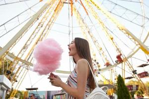 scatto all'aperto di donna dai capelli piuttosto lunghi in abito leggero e zaino bianco in posa sopra la ruota panoramica, in piedi con la bocca larga aperta e andando a mangiare zucchero filato foto