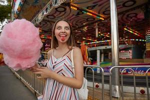 divertente donna bruna con gli occhi aperti con i capelli lunghi in abito estivo leggero in posa sopra il parco divertimenti, guardando la fotocamera felicemente e mostrando la lingua rosa dopo aver mangiato zucchero filato foto