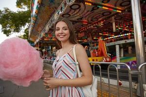 ritratto all'aperto di affascinante giovane donna in abito leggero con cinghie in piedi sopra il parco divertimenti in una calda giornata estiva, tenendo in mano zucchero filato e guardando la fotocamera con ampio sorriso foto