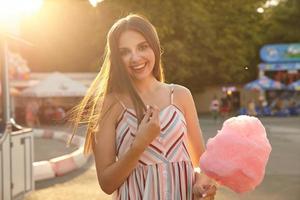 bella giovane donna dai capelli lunghi in abito estivo leggero con cinghie che guardano allegramente alla telecamera, toccandosi i capelli e sorridendo sinceramente alla telecamera, in piedi sopra il parco divertimenti in una giornata di sole foto