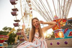 scatto all'aperto di gioiosa giovane donna graziosa con i capelli castani seduta sul parco divertimenti, sorridente ampiamente con gli occhi chiusi e alzando la mano con il gesto della vittoria, mangiando un cono gelato foto