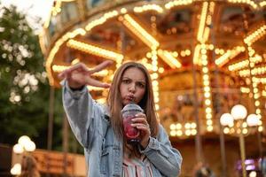 giovane bella donna dai capelli lunghi che fa una smorfia mentre beve limonata, guardando alla telecamera e accigliata, in piedi sopra la giostra con le dita sollevate nel gesto di vittoria foto