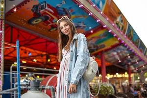 bella giovane donna dai capelli lunghi con occhiali da sole in testa in posa sopra le attrazioni del parco divertimenti, indossando un abito estivo romantico e un cappotto di jeans alla moda foto