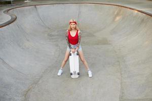 attraente donna bionda tatuata con i capelli lunghi in piedi sopra lo skate park in una calda giornata luminosa, indossando pantaloncini di jeans e top rosso, tenendo la tavola in mano foto