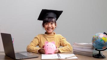 bambina asiatica che indossa un berretto da laurea con un salvadanaio rosa, risparmiando denaro, investendo il futuro, foto