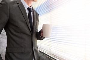primo piano di uomo d'affari che tiene una tazza di caffè mentre si trova davanti a una finestra in un ufficio foto