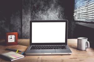 laptop con schermo bianco bianco e tazza di caffè, taccuino, penna e orologio sulla scrivania in legno con sfondo muro grunge.