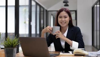 donna di affari asiatica sorridente che tiene una tazza di caffè e un computer portatile all'ufficio. guardando la telecamera. foto