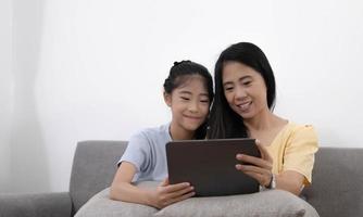 felice giovane mamma e figlia asiatiche che utilizzano tablet digitale, guardano video o navigano in Internet, si siedono sul divano a casa, copiano lo spazio. foto