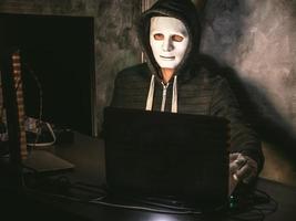 hacker di computer - uomo in maglietta con cappuccio con maschera che ruba i dati dal laptop foto