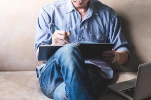 giovane casual che scrive alcuni dati nel taccuino con il laptop sul divano di casa. foto