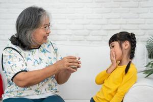 felice anziana nonna asiatica si siede accanto a sua nipote e nutre il latte fresco dal bicchiere per colazione a casa. concetto di una famiglia felice e si prende cura insieme, assistenza sanitaria in età prescolare foto