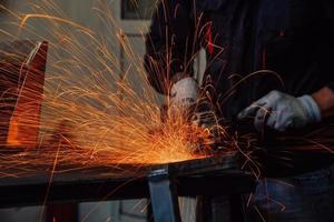 interno di una fabbrica di ingegneria dell'industria pesante con un lavoratore industriale che utilizza una smerigliatrice angolare e taglia un tubo di metallo. appaltatore in uniforme di sicurezza e elmetto per la produzione di strutture metalliche. foto