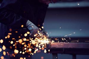 interno di una fabbrica di ingegneria dell'industria pesante con un lavoratore industriale che utilizza una smerigliatrice angolare e taglia un tubo di metallo. appaltatore in uniforme di sicurezza e elmetto per la produzione di strutture metalliche. foto