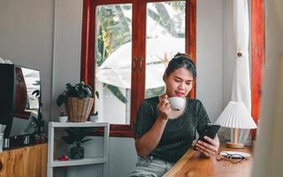 donna asiatica con un bel sorriso che guarda sul telefono cellulare durante il riposo in una caffetteria, donna tailandese felice si siede al bancone bar in legno bevendo caffè rilassante nella caffetteria durante il tempo libero foto