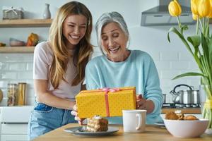 donna anziana sorpresa che riceve una confezione regalo da sua figlia mentre trascorre del tempo in cucina insieme foto