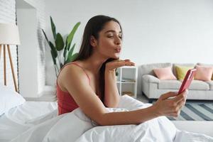 attraente giovane donna in possesso di smart phone e soffiando un bacio mentre si siede a letto a casa foto