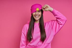 felice giovane donna in camicia con cappuccio che regola il suo cappello funky su sfondo rosa foto