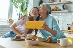 donna anziana felice che riceve una confezione regalo da sua figlia adulta mentre entrambi sono seduti in cucina foto