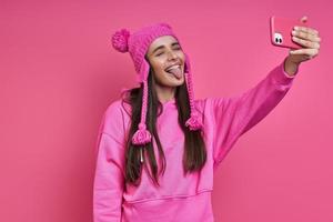 giovane donna giocosa con cappello funky che fa selfie e fa una smorfia su sfondo rosa foto