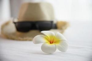 cappello di roba turistica occhiali da sole e fiore di plumeria in camera da letto bianca - felice vacanza relax vacanza e concetto di hotel foto