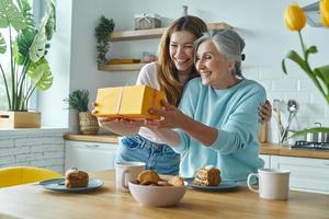 madre anziana sorpresa che riceve una confezione regalo da sua figlia mentre era seduta in cucina insieme foto