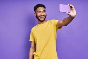 uomo africano felice che fa selfie con lo smartphone mentre si trova su sfondo viola foto