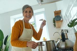 donna anziana felice che guarda l'obbiettivo mentre cucina nella cucina domestica foto