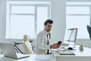 uomo sicuro di sé in camicia e cravatta guardando il suo smartphone mentre è seduto in ufficio foto