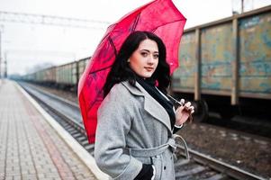 ragazza bruna in cappotto grigio con ombrello rosso nella stazione ferroviaria. foto