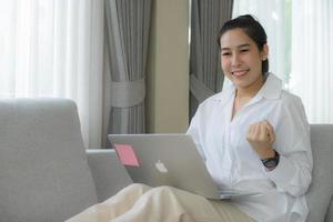 allegra giovane ragazza asiatica seduta sul divano a casa con un computer portatile e festeggia il successo, sorridendo ampiamente felice, positiva, sicura di sé con entrambe le braccia in alto. foto