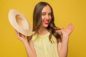 la ragazza affascinante e divertente con le labbra rosa sta rinunciando alla fotocamera mentre tiene il cappello e posa alla fotocamera su sfondo giallo foto