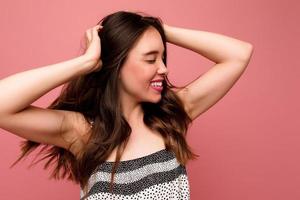 donna divertente uscita con i capelli scuri che indossa un abito estivo luminoso ballando e divertendosi in studio su sfondo rosa foto