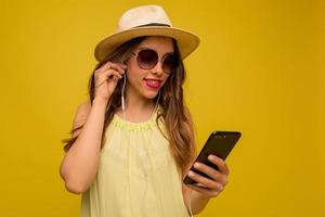 il ritratto al chiuso di una ragazza alla moda con i capelli lunghi e le labbra rosa che indossa un cappello e un vestito giallo con gli occhiali da sole sta ascoltando musica e guardando lo smartphone su sfondo giallo foto