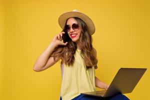 felice ragazza adorabile con i capelli mossi che indossa un vestito giallo in occhiali da sole e cappello con labbra rosa sta guardando lontano mentre parla al telefono e tiene in mano il laptop foto