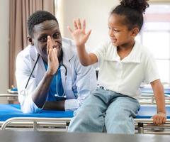 paziente bambino seduto sul letto d'ospedale con un medico africano per cure mediche. bambina sorridente felice in buona salute dopo il controllo professionale in clinica. medico, pediatra che controlla, esamina il bambino foto