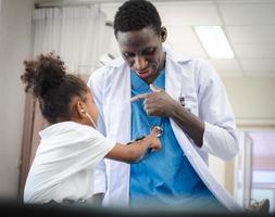 messa a fuoco selettiva al medico persona che gioca con un simpatico bambino afro paziente nel reparto ospedaliero. amichevole pediatra divertente ragazza ragazzo godendo con lo stetoscopio per il battito cardiaco durante l'esame medico. foto