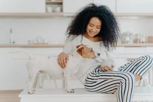 inquadratura di una giovane donna dall'aspetto piacevole concentrata, gioca con il cane preferito, tiene una tazza di bevanda, posa insieme in cucina, esprime amore, ha tempo libero. rapporto tra persone e animali foto