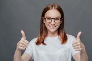 felice donna positiva con un tenero sorriso sul viso, ha i capelli castani, alza due pollici, dimostra la sua approvazione, isolata su sfondo grigio, indossa occhiali, indossa una maglietta bianca casual. linguaggio del corpo foto