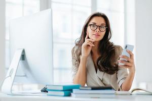 L'impiegata dell'azienda in abbigliamento formale utilizza gadget moderni per il lavoro, posa sul posto di lavoro vicino al monitor, pianifica l'orario di lavoro, sfoglia le informazioni online, posa contro la finestra. orario di lavoro foto