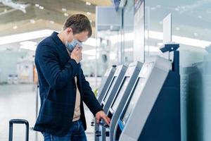 inquadratura del passeggero uomo posa vicino al banco del check-in in aeroporto, ha un servizio self-service, indossa una maschera medica per evitare la diffusione di malattie respiratorie durante l'epidemia di coronavirus, viaggia e vuole volare all'estero foto