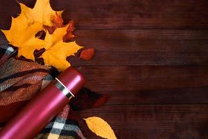 disposizione piatta autunnale. thermos con caffè caldo, plaid e foglie cadute su fondo di legno. inizio della stagione autunnale. foto