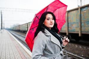 ragazza bruna in cappotto grigio con ombrello rosso nella stazione ferroviaria. foto